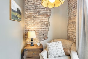 A cozy room with a brick wall, a wooden nightstand with a lamp, a clock, and a patterned sofa. - On Dtwn Square! Chic Lawrenceburg Condo w/ Balcony (Lawrenceburg)