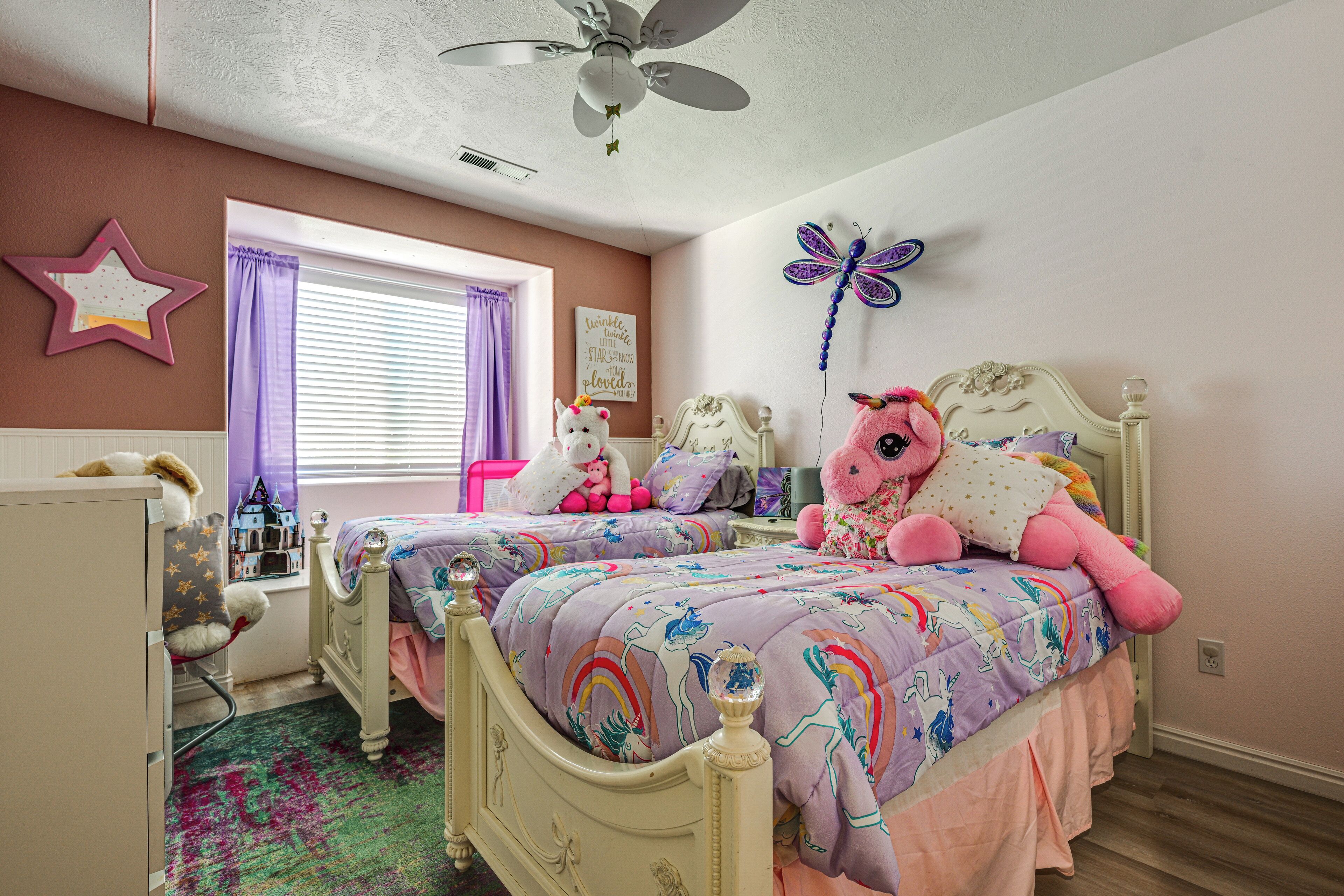 A bedroom with two beds, a ceiling fan, a star decoration, a butterfly wall art, and a window with purple curtains.