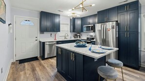 A modern kitchen with dark cabinets, stainless steel appliances, a central island, and a unique chandelier. - Coastal Serenity-3BR Luxury Home in Cherry Grove (North Myrtle Beach)