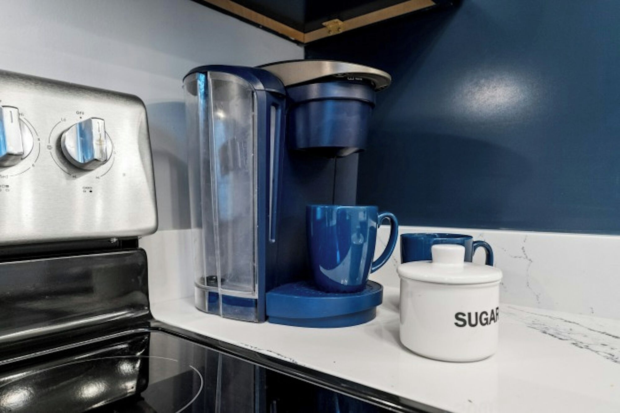 Cuina amb una cafetera, dues tasses blaves i una tassa blanca amb la paraula 'SUGAR'. 