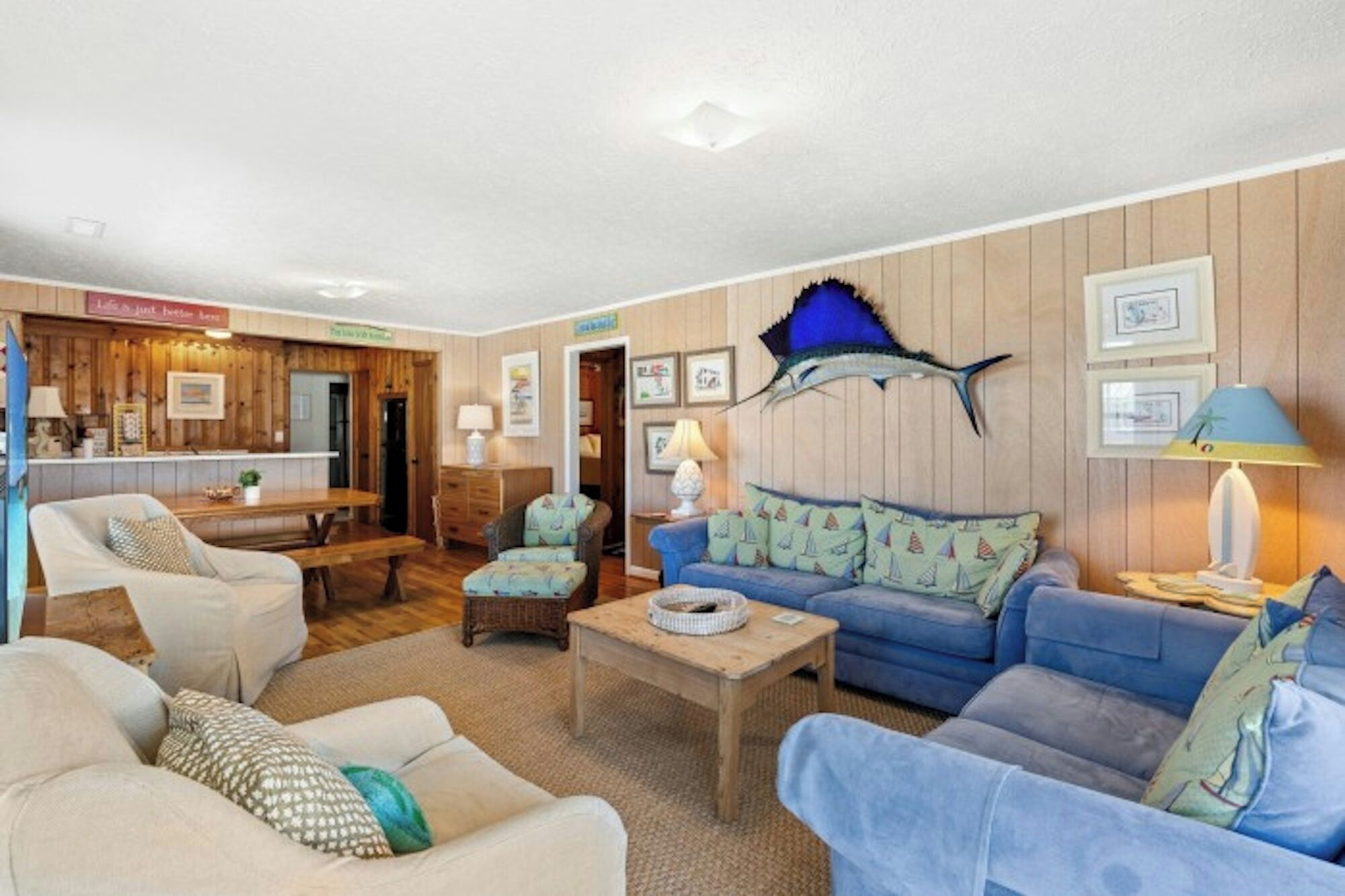 A cozy living room with a wooden wall, a blue sofa, a dining table, and a large fish mounted on the wall.