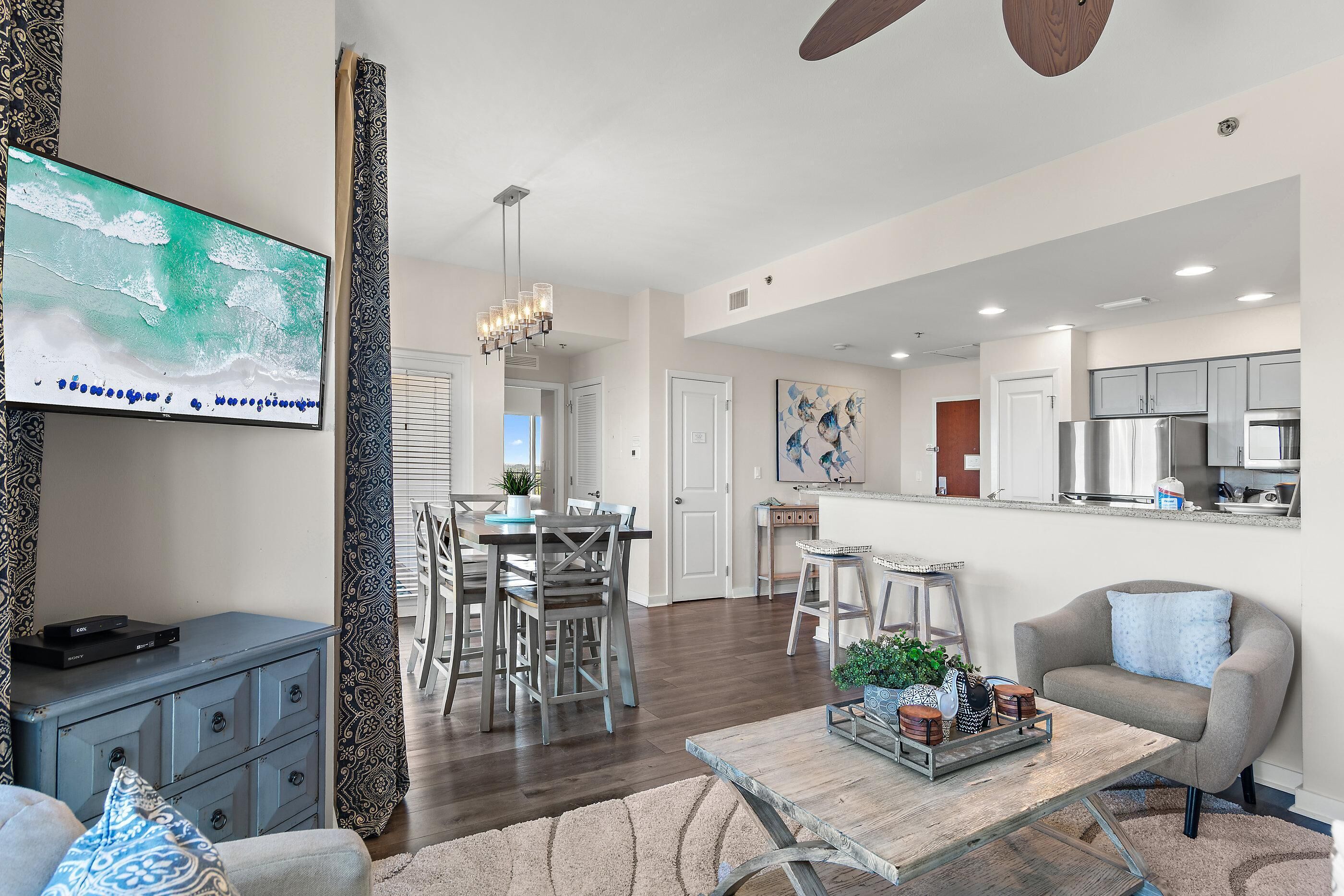 A modern living room with a flat-screen TV, a dining area with a chandelier, a kitchen with stainless steel appliances, and a gray armchair.