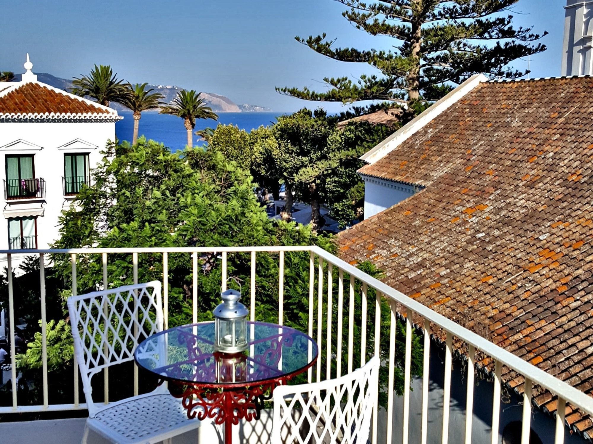 Ein Balkon mit Glastisch und Stühlen, der auf eine Küstenlandschaft mit Gebäuden und Bäumen blickt.