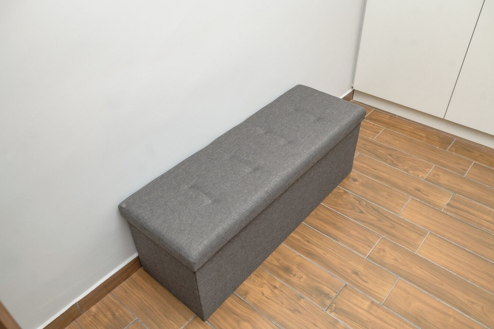 A grey bench in a corner of a room with wooden flooring and white walls.