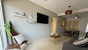Una sala de estar con un sofá, una silla, un televisor montado en la pared y un comedor con mesa y sillas.