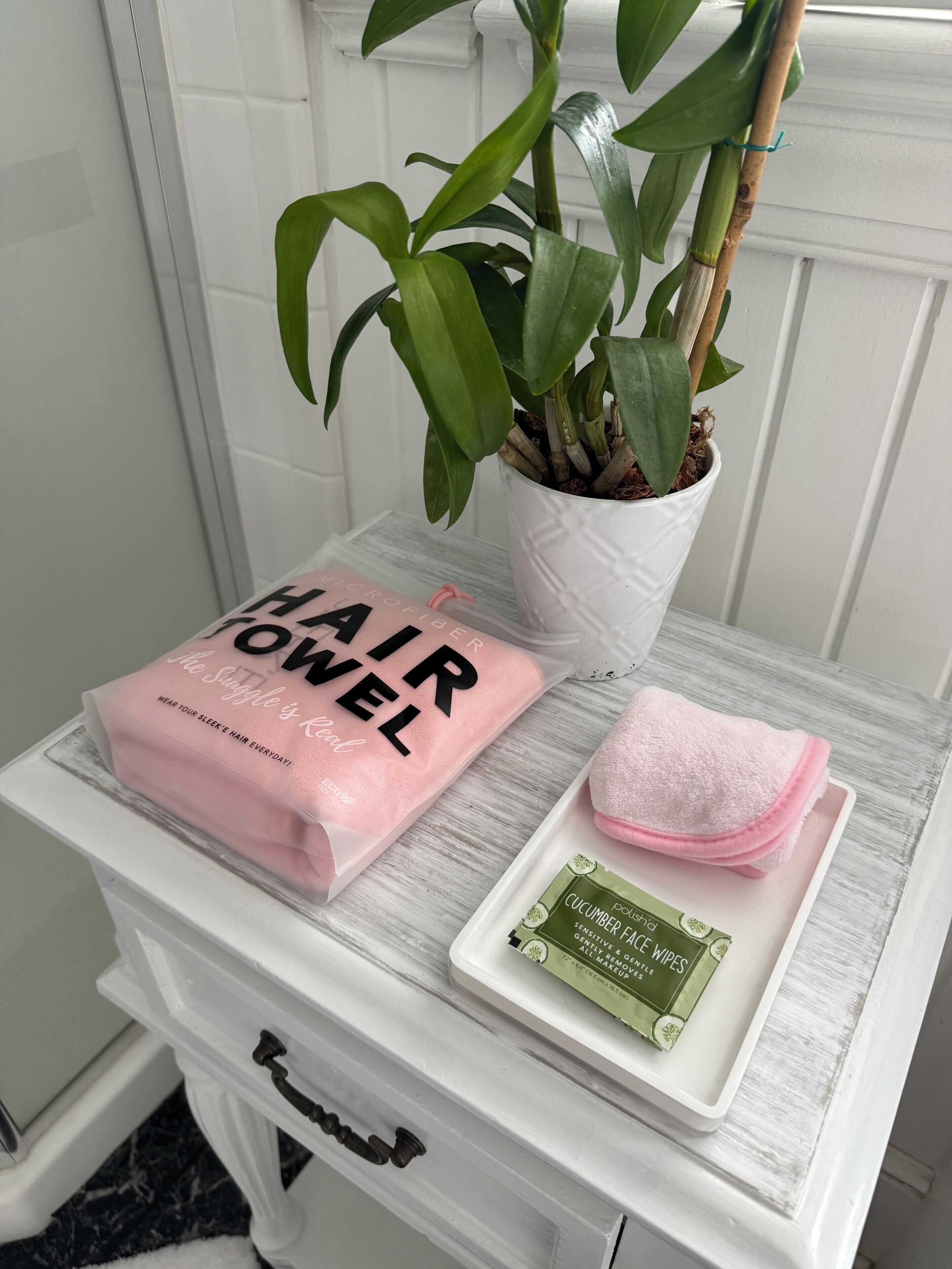 Un meuble-lavabo de salle de bain avec un meuble blanc, une plante en pot, une serviette rose et un produit vert posé sur le plateau.