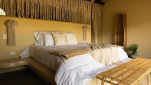 A bed with white bedding, a woven bench, and macramé wall decor. - Holistic EcoHotel (San Nicolás)