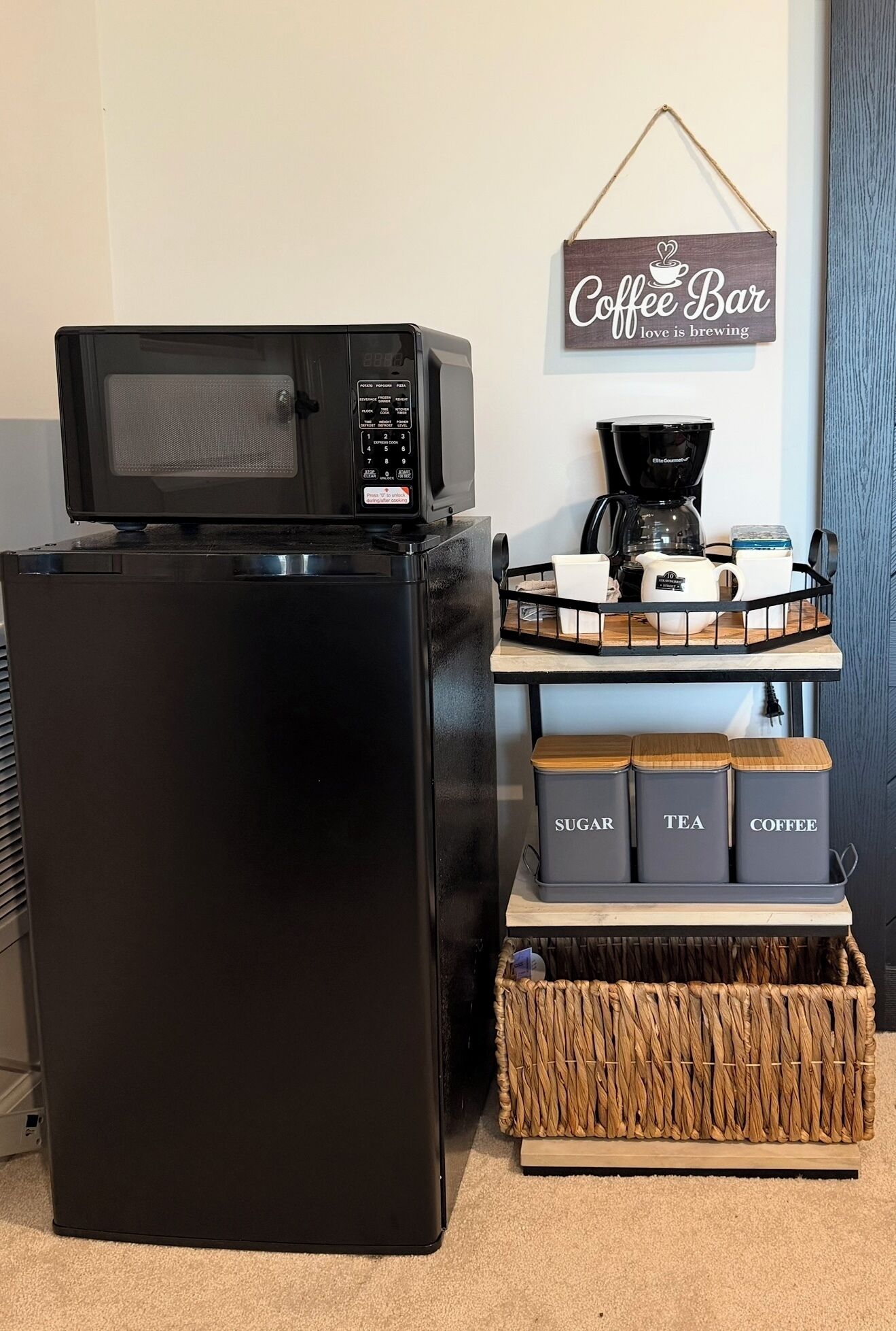 Un réfrigérateur noir avec un four à micro-ondes sur le dessus, une machine à café et des récipients étiquetés pour le sucre, le thé et le café.