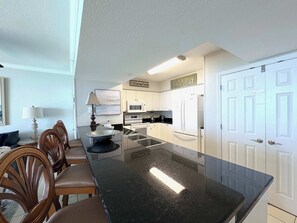 A kitchen with white cabinets, a black countertop, a dining table with chairs, a microwave, a refrigerator, and a 'blessed' sign above the cabinets. - Oceanfront, 23rd floor Condo, Pools, Gym, Wifi (Panama City Beach)