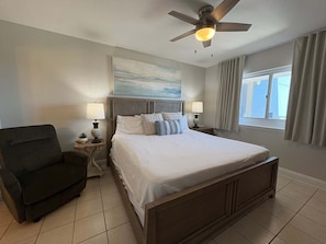 A bedroom with a bed, two bedside tables with lamps, a chair, a ceiling fan, a window with curtains, and a painting on the wall. - Oceanfront, 23rd floor Condo, Pools, Gym, Wifi (Panama City Beach)
