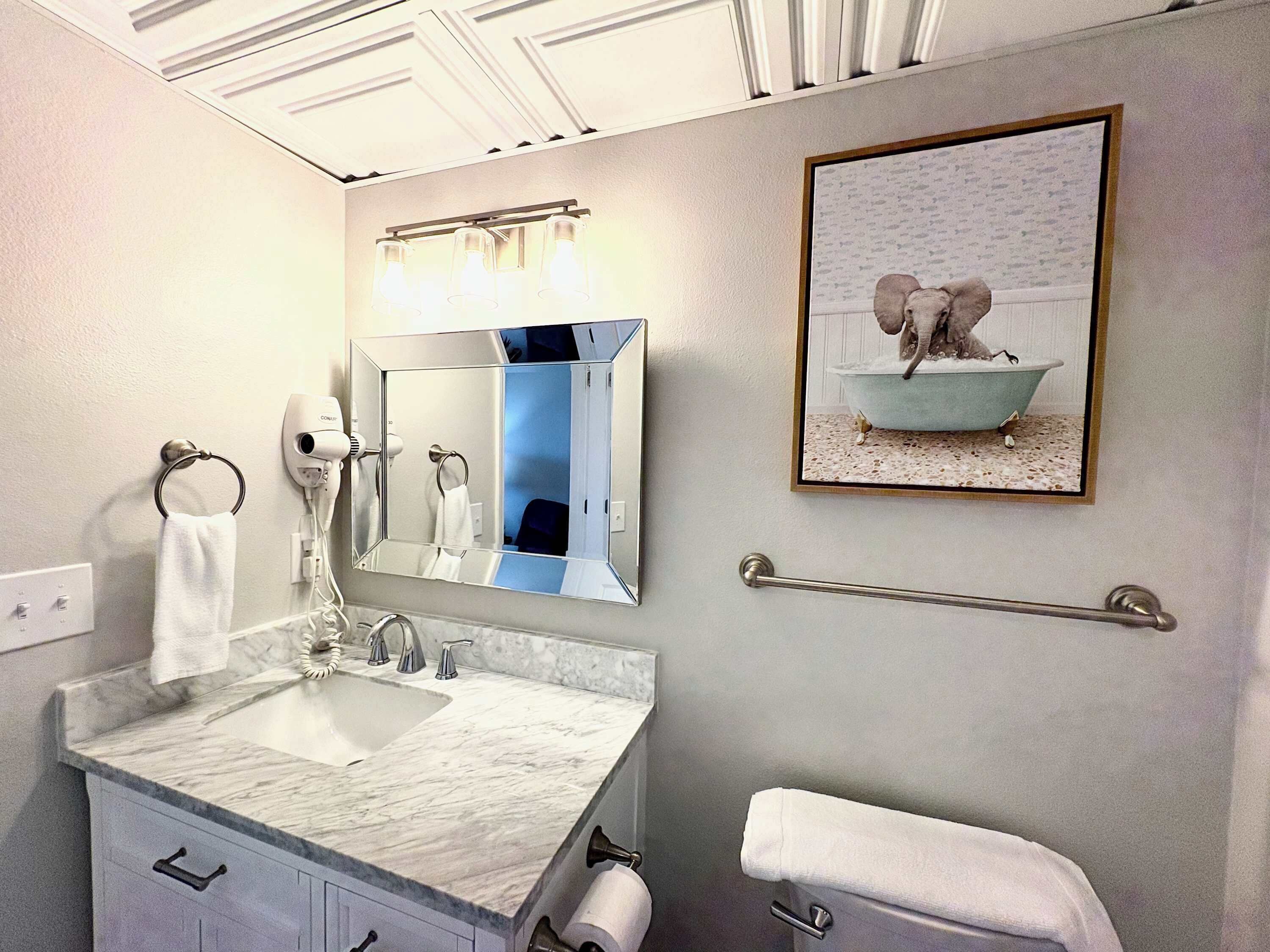 A bathroom with a sink, mirror, towel rack, hairdryer, and a framed picture of a bathtub.