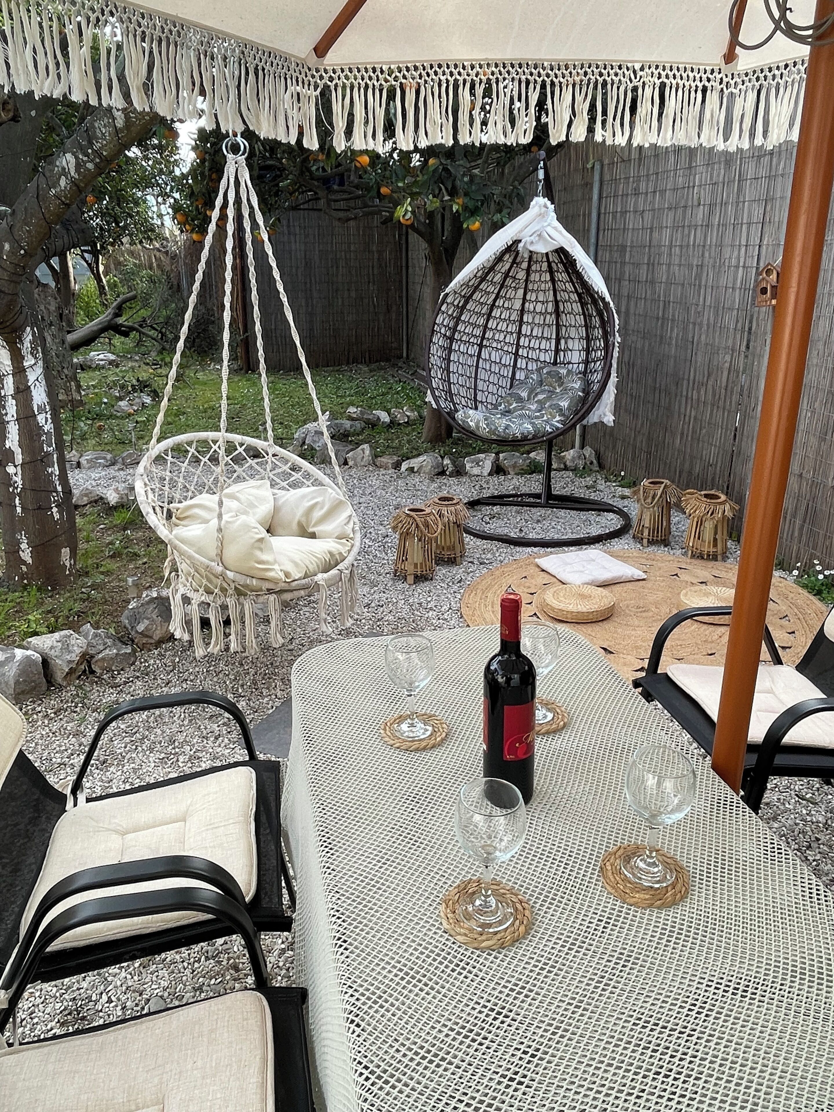 An outdoor seating area with a table, chairs, a hanging chair, and a bottle of wine.