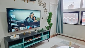 Ett modernt vardagsrum med en stor platt-TV, en turkosfärgad möbel för underhållningsutrustning, ett runt soffbord med en vas och utsikt över staden genom ett fönster.