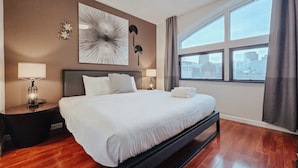 Ett hotellrum med en stor säng, två sängbord med lampor, en modern, abstrakt målning och utsikt över staden genom stora fönster.
