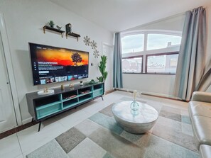 Ein Wohnzimmer mit Flachbildfernseher, Couchtisch, Sofa, einem Fenster mit Vorhängen und einer Pflanze.