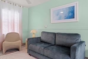 Een kamer met een grijze bank, een rieten stoel, een lamp en een schilderij aan de muur.