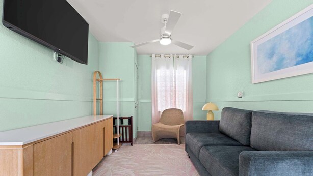 A living room with a flat-screen TV mounted on the wall, a ceiling fan, a sofa, a chair, and a small table. - Handicap Accessible Suite in Clearwater Beach (Clearwater)