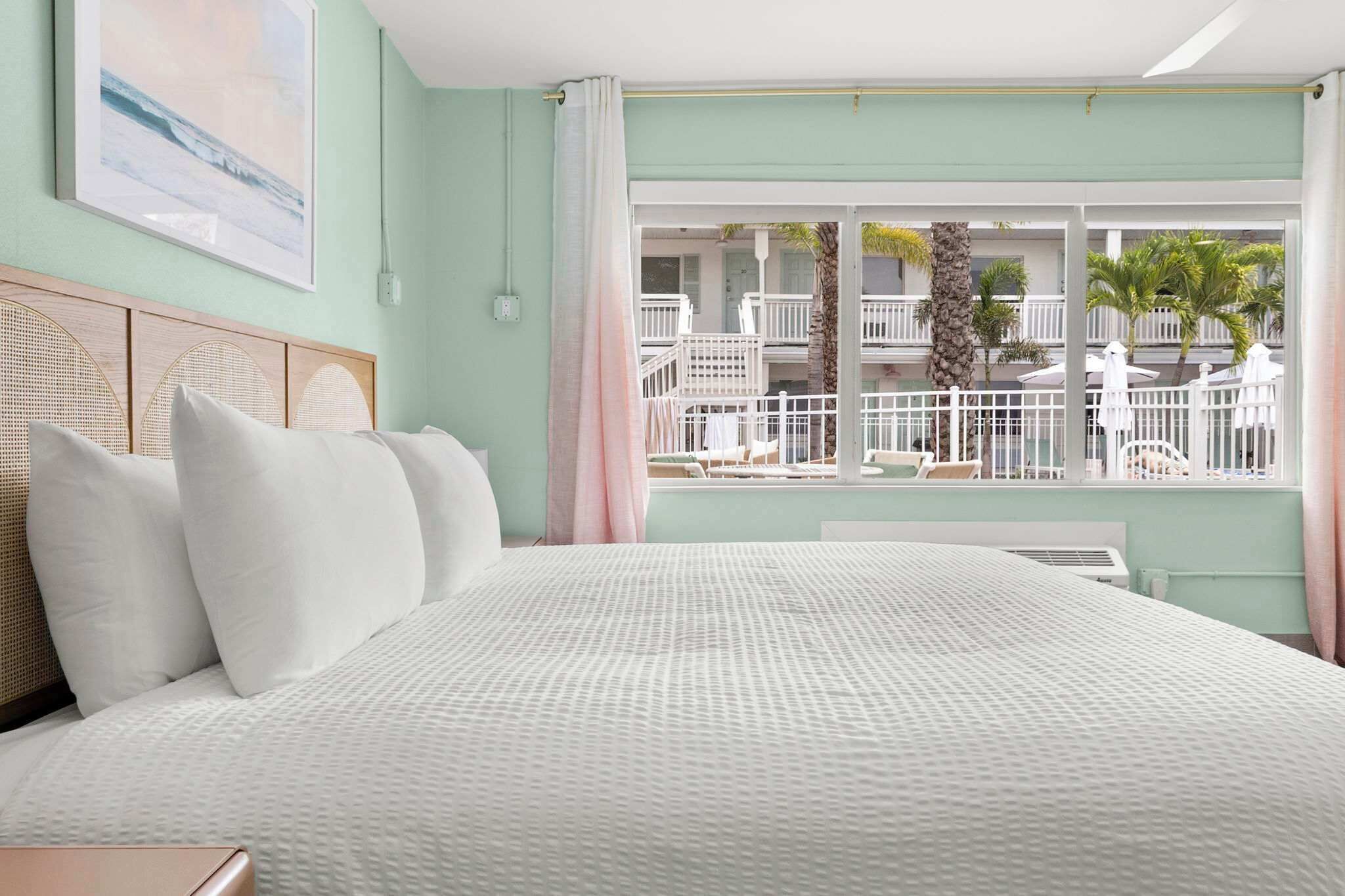 A hotel room with a large bed, a view of a balcony with outdoor furniture, and a painting on the wall.