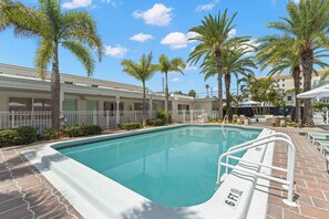Una piscina con muelle blanco y un cartel de prohibido bucear, rodeada de palmeras y un edificio con fachada blanca.