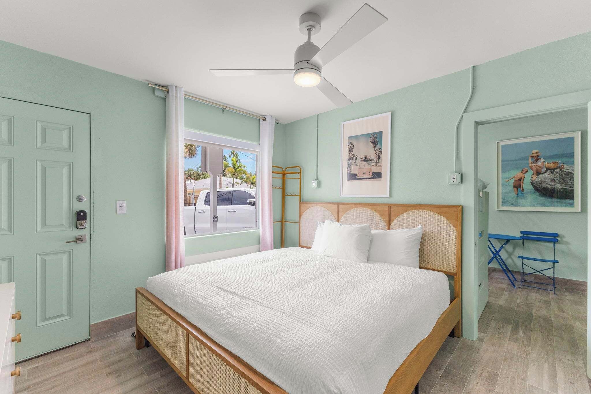 A bedroom with a bed, a ceiling fan, a window with a view of the street, and a painting on the wall.