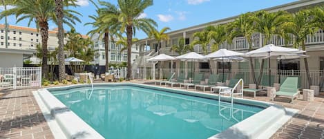 Una piscina con sillas de descanso y sombrillas, rodeada de palmeras y un edificio con balcones.