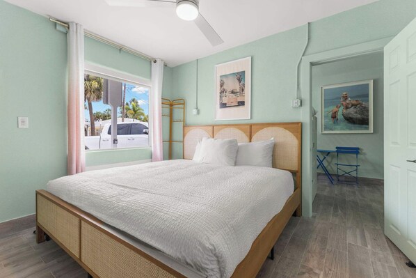 Un dormitorio con cama, ventana con vista a palmeras y puerta que da a otra habitación.