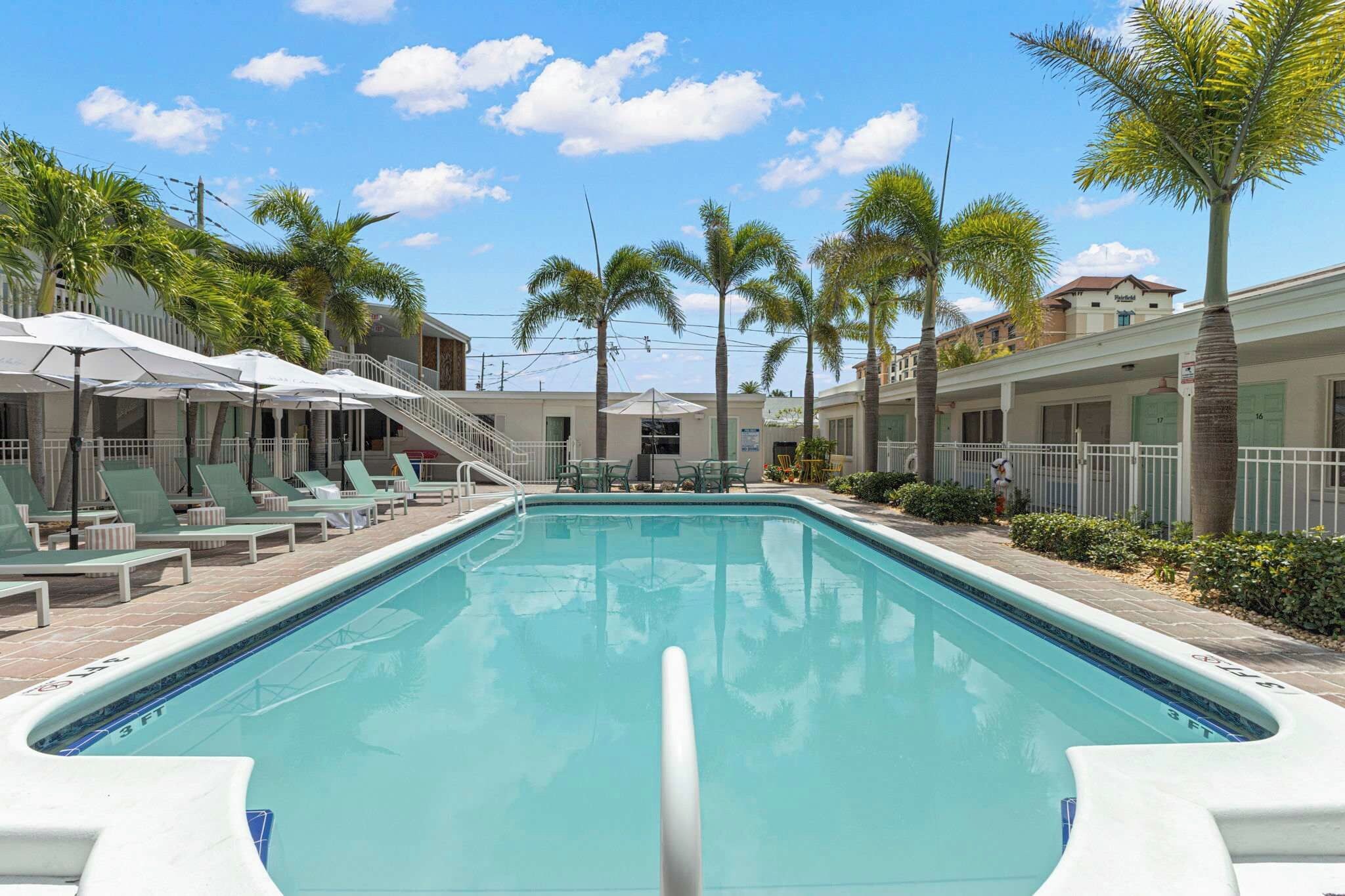Una piscina con sillones de playa y sombrillas, rodeada de palmeras y edificios.