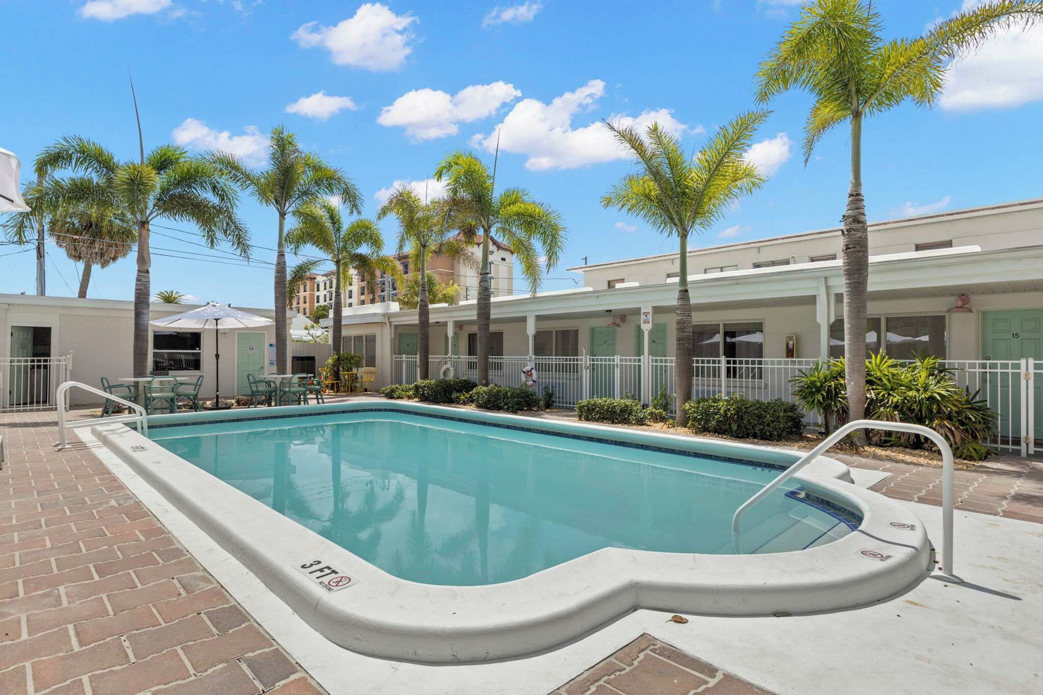 A swimming pool with a 3ft depth marker, surrounded by palm trees and a building with a white facade.