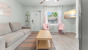 Un salon comprenant un canapé, une table en bois, deux fauteuils roses, un ventilateur de plafond, une planche de surf, une porte et une fenêtre donnant sur des palmiers.