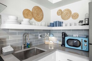 A modern kitchen with grey tiles, a sink, a blue microwave, and decorative woven wall art. - NEW! Modern King Suite | Steps to the Beach! (Clearwater)