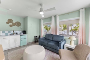 A modern living room with a sofa, a round coffee table, a wicker chair, a small side table with a lamp, and a kitchen area with cabinets and a coffee maker. - Coastal Suite - Sparkling Pool, Steps to the Beach (Clearwater)