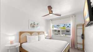Quarto com cama, mesas de cabeceira, ventilador de teto, janela com cortinas, televisão e prateleira fixada na parede.