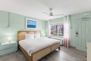 A bedroom with a bed, bedside tables, a ceiling fan, a window with curtains, and a door. - The Cabana Suite | Steps to the Beach! (Clearwater)