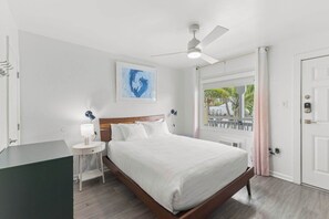 A hotel room with a bed, bedside table, lamp, ceiling fan, artwork, window with curtains, and a door. - NEW! North Petite Room | Steps to the Beach! (Clearwater)