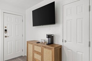 A room with a white door, a mounted flat-screen TV, a coffee machine, and a wooden cabinet with drawers. - NEW! North Petite Room | Steps to the Beach! (Clearwater)