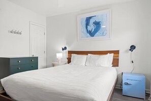 Un dormitorio ordenado con cama, mesitas de noche, cómoda, cuadro en la pared y un refrigerador pequeño.