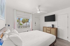 Ein Schlafzimmer mit Bett, Deckenventilator, Fernseher, Kleiderschrank und Fenster mit Blick auf Palmen.