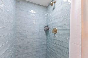 Ducha en esquina con azulejos azul claro, cabezal de ducha dorado y toallero blanco con botellas.