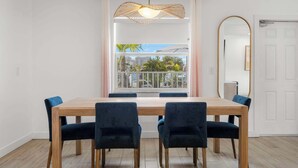 Een eethoek met een houten tafel en donkere stoelen, een hangende lamp, een groot raam met uitzicht op gebouwen en palmbomen, en een witte deur met een spiegel.