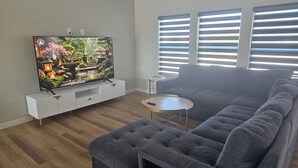 A living room with a flat-screen TV, grey sofa, ottoman, and a small round table.