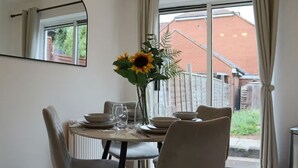 A dining area with a table set for four, a vase with sunflowers, and a view of a brick building through a window. - Central 3 Bed Home, Parking Garden Milton Keynes (Bradwell Common)
