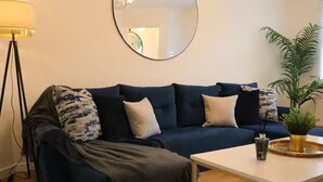 A living room with a dark blue sofa, a round mirror, a floor lamp, a coffee table, and a potted plant. - Central 3 Bed Home, Parking Garden Milton Keynes (Bradwell Common)