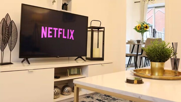 A living room with a TV displaying 'NETFLIX,' a white entertainment unit, a table with a tray, a potted plant, and a window with a view. - Central 3 Bed Home, Parking Garden Milton Keynes (Bradwell Common)