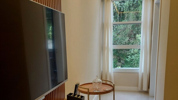 A modern hotel room with a flat-screen TV, a wooden desk, a small round table, and a large window with curtains. - Quiet apartment, excellent location in Copacabana, 5 minutes from the beach. (Rio de Janeiro)