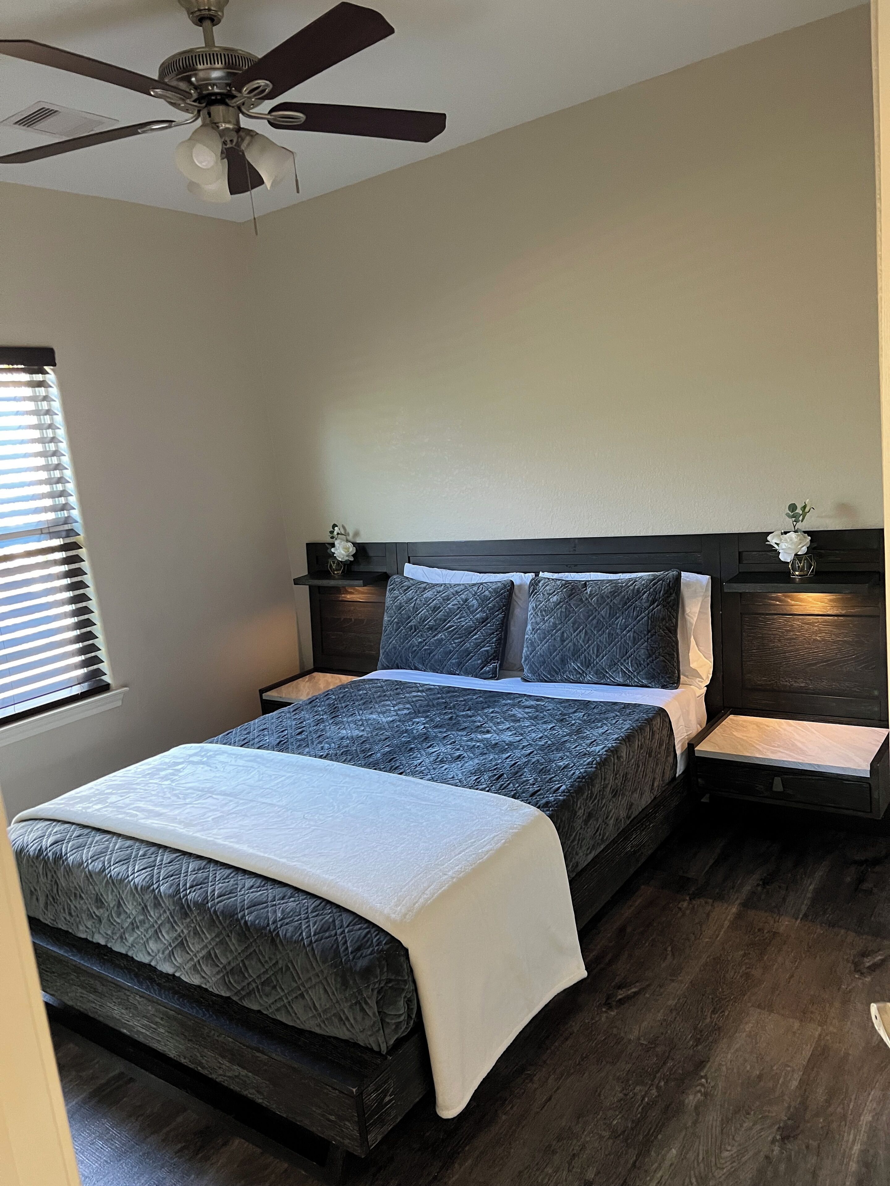 Une chambre à coucher avec un lit, des tables de chevet, un ventilateur de plafond et une fenêtre avec des stores.