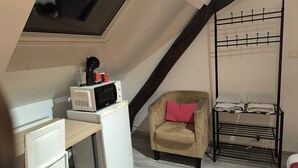 A compact room with a loft bed, a small desk with a microwave, a chair, and a metal drying rack.