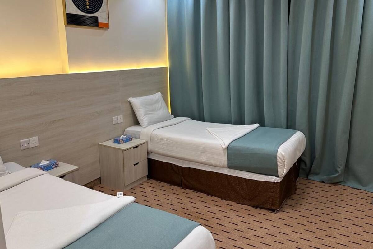 A hotel room with two beds, a bedside table with a lamp, a wall-mounted air conditioner, and a window with curtains.