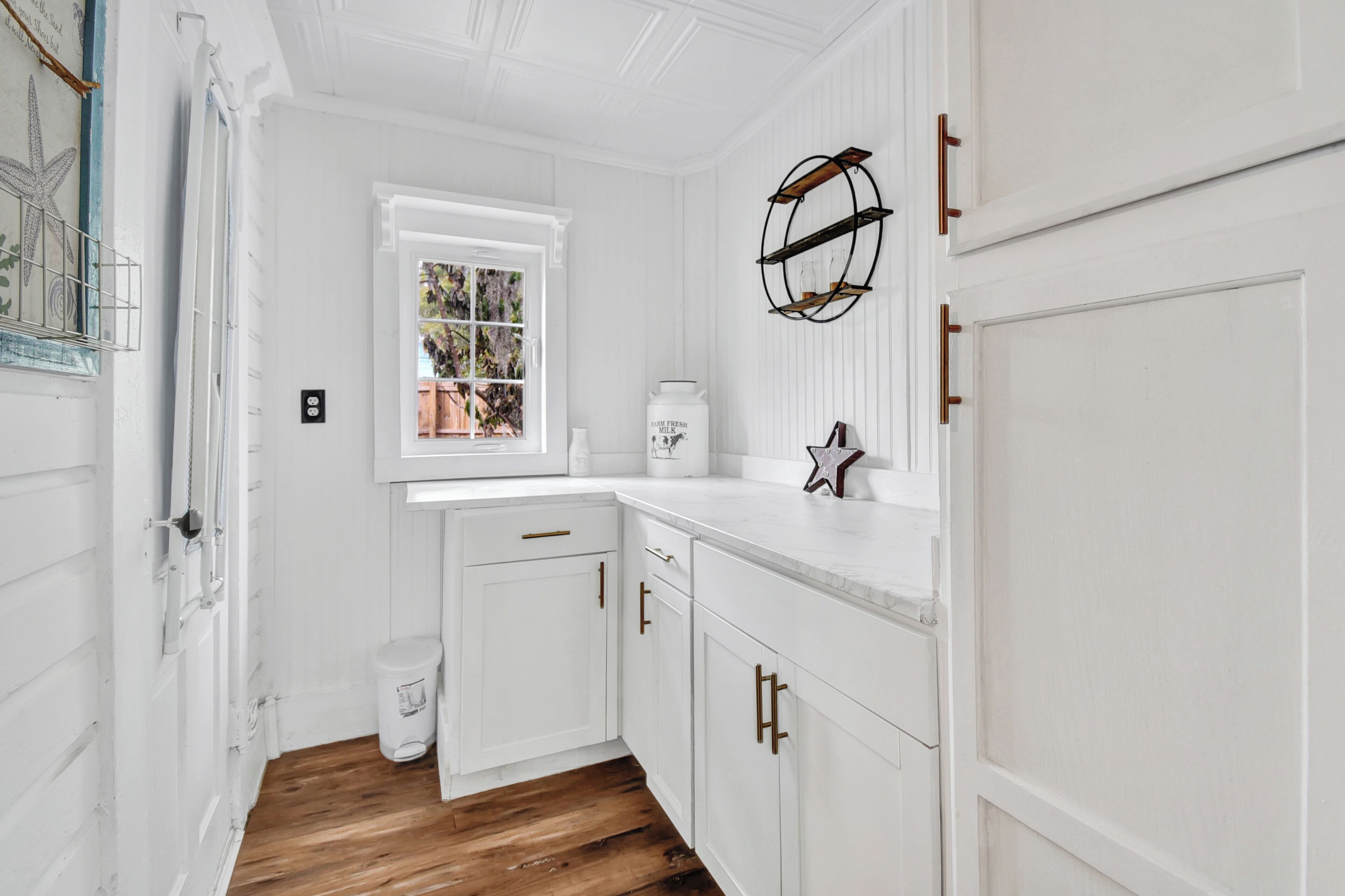 A white kitchen with wooden flooring, white cabinets, a window, and a decorative metal shelf.