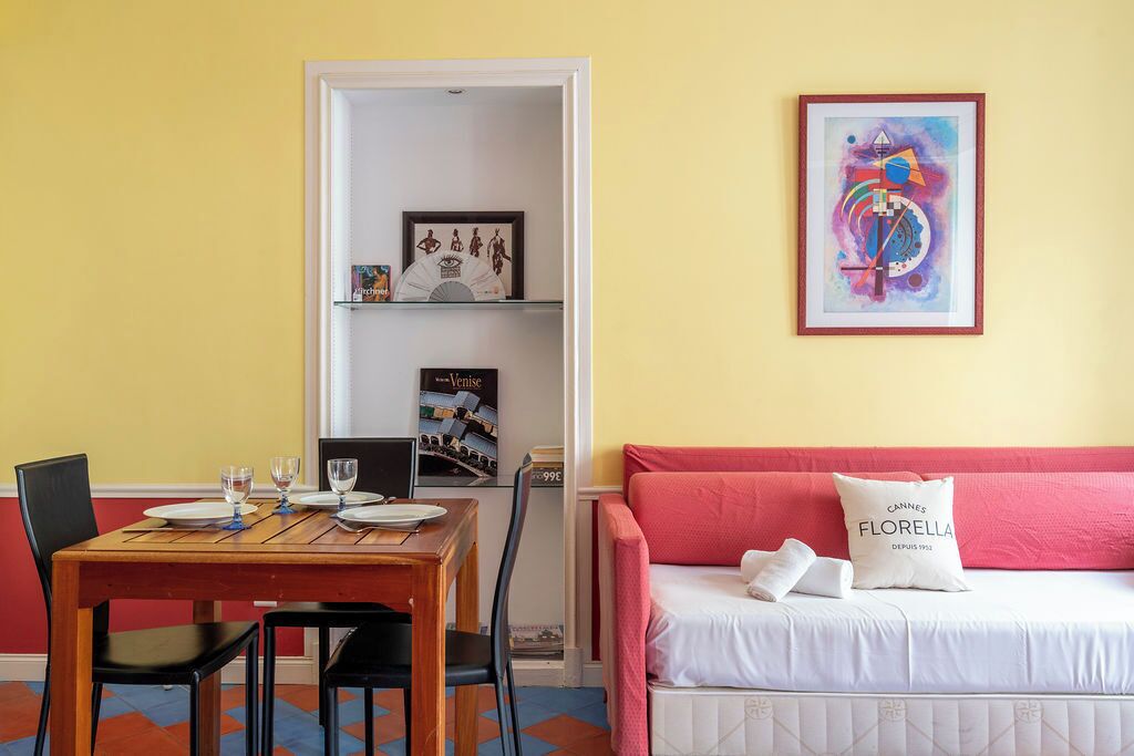 A room with a dining table set for two, a bed with a pink headboard, and a framed abstract painting on the wall.