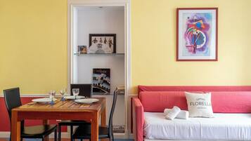 A room with a dining table set for two, a bed with a pink headboard, and a framed abstract painting on the wall.